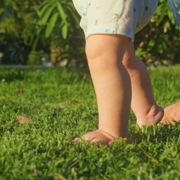 . Baby learns to walk with the help of his mother on a green grass in the park.